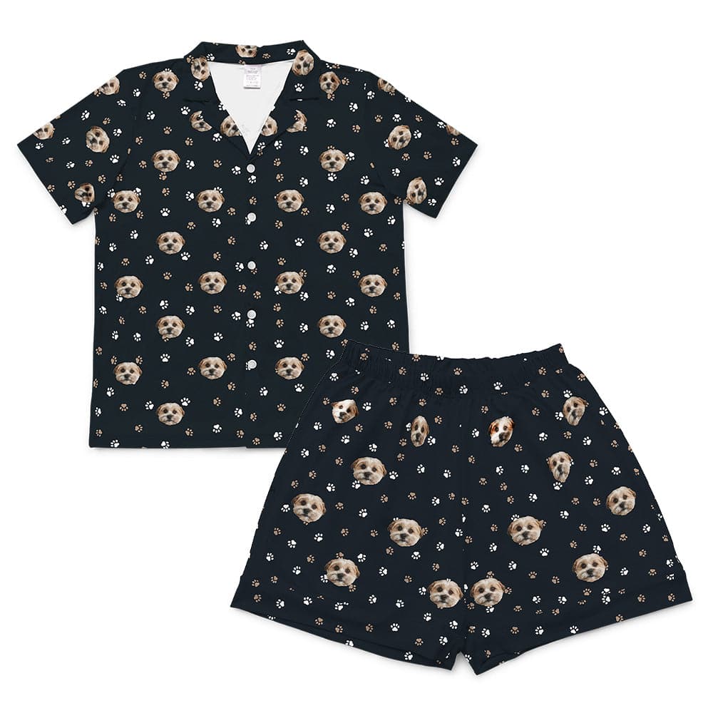 A black short-sleeve pajama set, consisting of a button-down top and matching short-length shorts. Both pieces feature a repeating pattern of light brown and white dog faces interspersed with scattered white and light brown paw prints. 