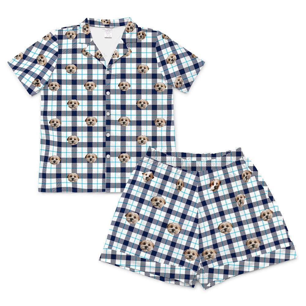 A short-sleeve pajama set, consisting of a button-down top and matching short-length shorts. Both pieces feature a white background with a large navy blue and thin light blue plaid pattern, and a repeating pattern of light brown and white dog faces interspersed across the plaid.