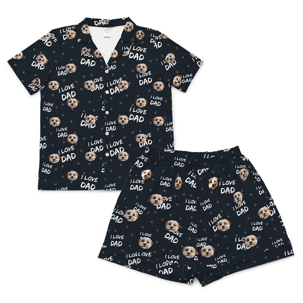 A black short-sleeve pajama set, consisting of a button-down top and matching short-length shorts. Both pieces feature a repeating pattern of light brown and white dog faces, white text reading "I LOVE DAD", and small white and light brown paw prints.