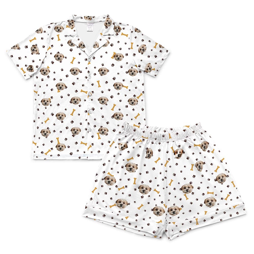 White short-sleeve pajama set, including a button-down top and matching shorts, both featuring a scattered pattern of dog faces, brown paw prints, and yellow-brown dog bones.