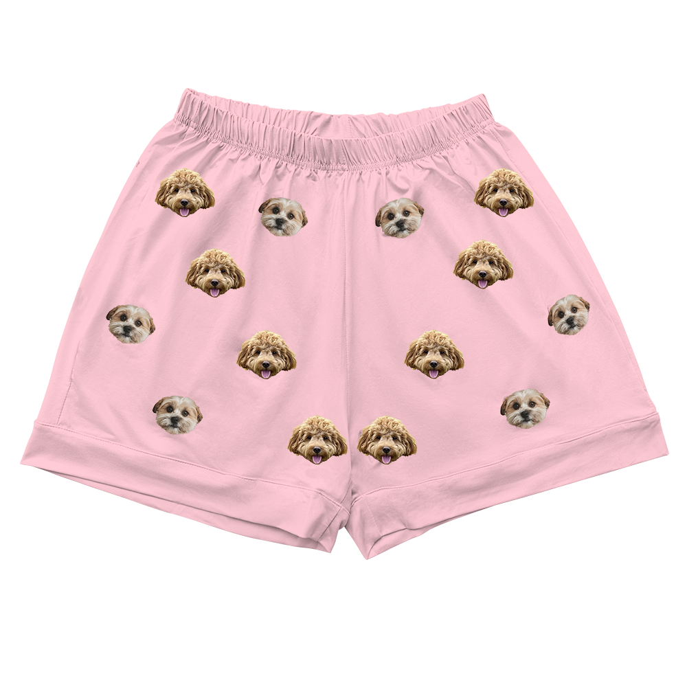 Pink pajama shorts with elastic waistband and rolled cuffs, featuring a scattered print pattern of realistic doodle dog faces in brown, tan, and cream colors.