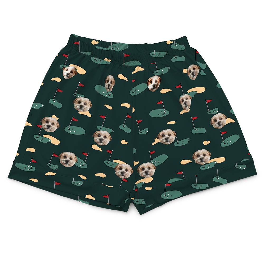 Dark green pajama shorts with an elasticated waist. The shorts are covered in a repeating golf-themed pattern featuring alternating elements: cartoon depictions of golf putting greens with a red flag, yellowish sand traps, and smiling headshots of a dog.