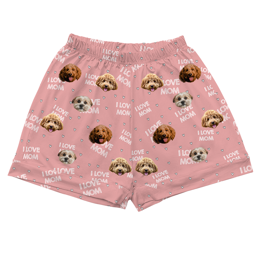 A pair of pink, short-length pajama shorts featuring a repeating pattern of various dog faces in different shades of brown and white, and white text reading "I LOVE MOM" interspersed with small white hearts. 