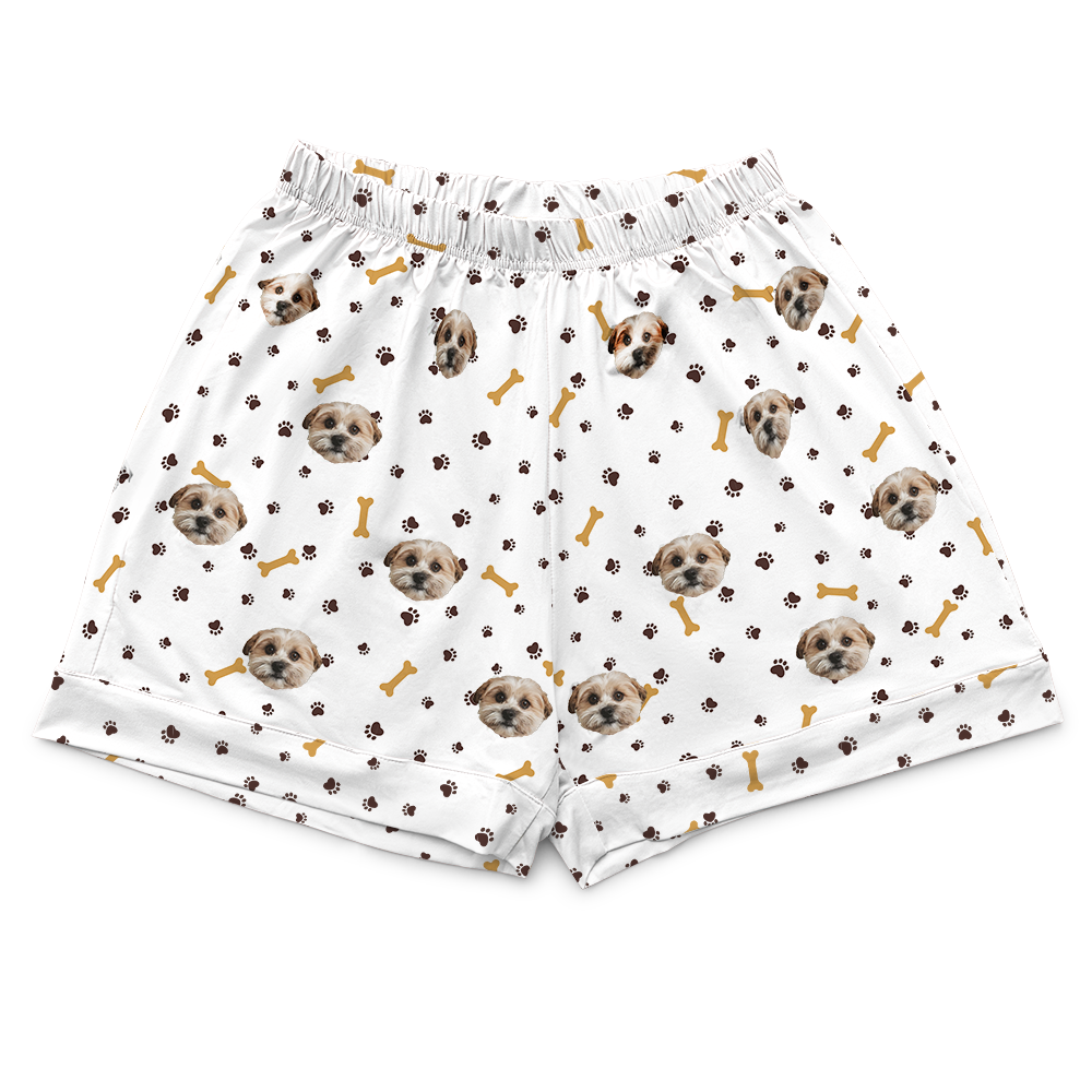 A pair of white, elastic-waist pajama shorts with a repeated pattern of small illustrations. The pattern includes dog faces (specifically, a light brown Poodle or similar small breed), dark brown paw prints, and light brown bones, all scattered evenly across the fabric.