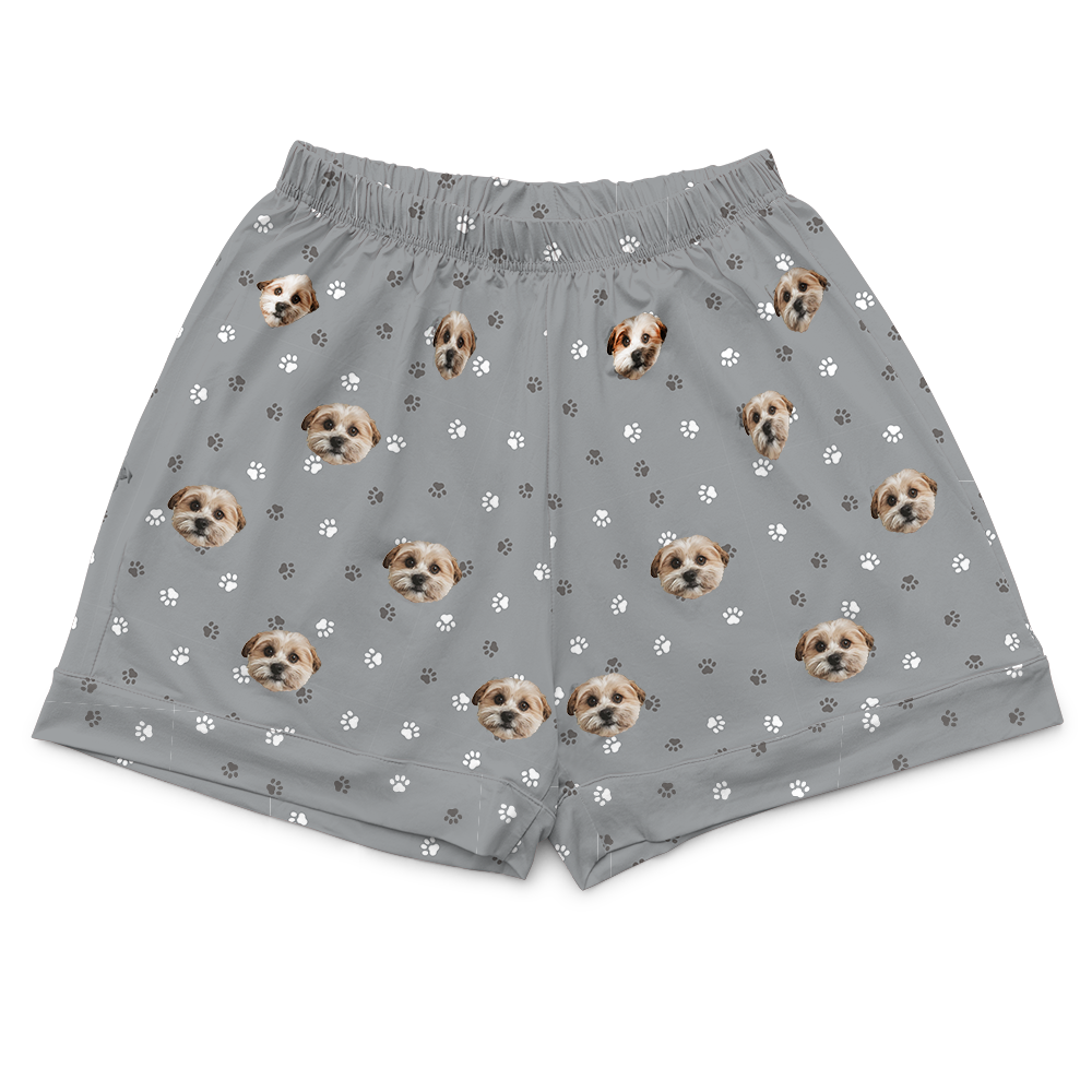 A pair of slate grey pajama shorts with a scattered pattern of small white and dark grey paw prints and multiple repeated images of a light brown and white dog's face. 