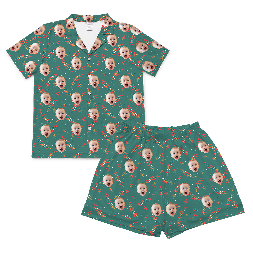 A two-piece set of short-sleeve pajamas in a green color with a pattern of red berries and green foliage. The pattern is customized with multiple small, repeating images of a baby's smiling face with its mouth open, covering the entire fabric of both the shirt and the shorts. 