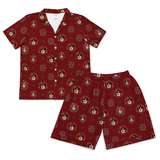 A two-piece pajama set featuring a short-sleeve button-up shirt and matching shorts in a green plaid pattern with Dalmatian dog faces, pink figures, and small colorful patches, shown on a plain white background.