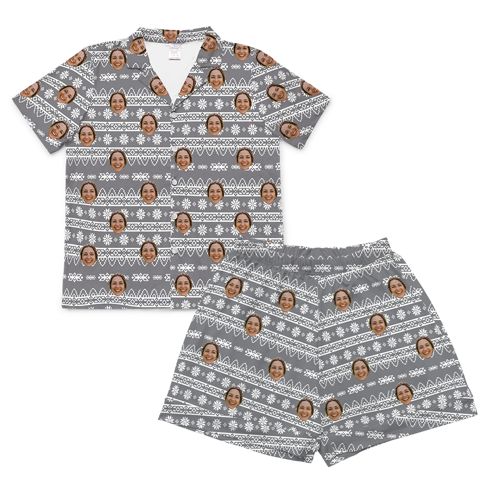 A matching short-sleeved pajama set, consisting of a button-up shirt and shorts, laid flat against a solid light gray background. Both pieces feature a gray background patterned with a white, Nordic-style fair isle design, including stripes, crosses, and snowflakes, resembling a holiday sweater pattern.