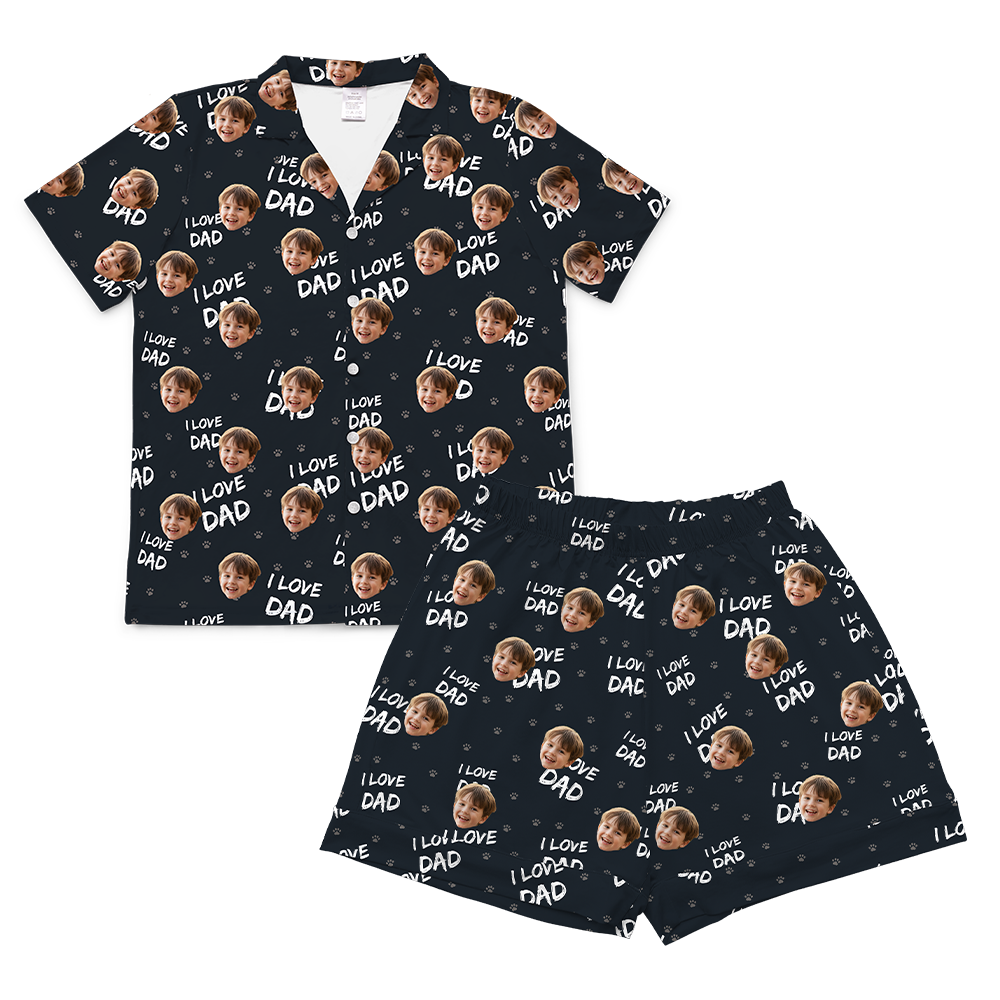 A two-piece set of short-sleeve pajamas in a black color. The pajamas are customized with a repeating pattern that includes the text "I LOVE DAD" in white, small images of white paw prints, and small, repeating images of a young child's smiling face.