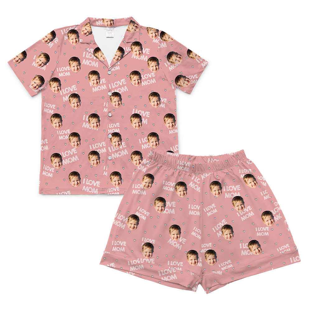 A two-piece set of short-sleeve pajamas in a solid light pink color. The pajamas are customized with a repeating pattern that includes the text "I LOVE MOM" and small heart shapes in white, along with small, repeating images of a young child's smiling face.