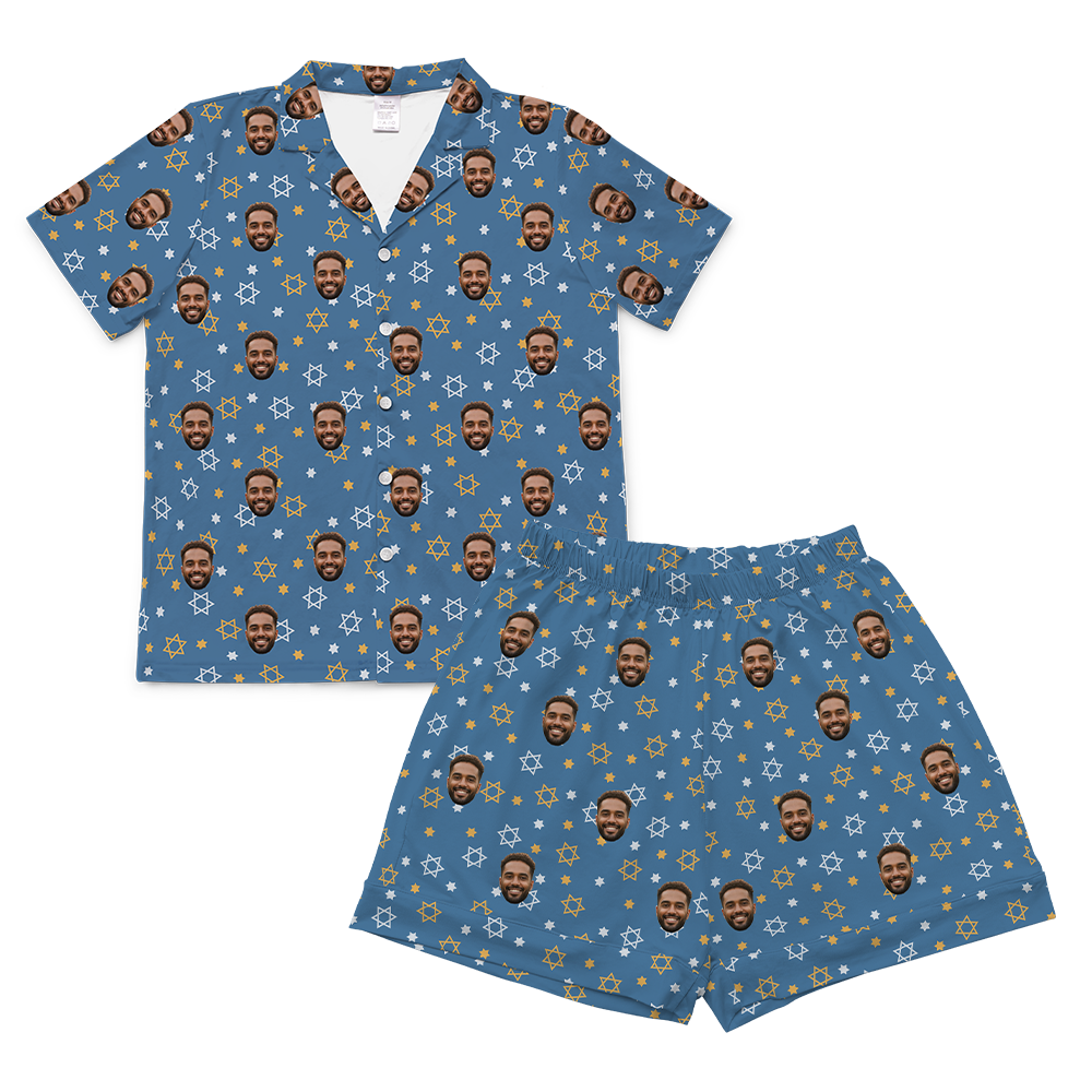 A matching short-sleeved pajama set, consisting of a button-up shirt and shorts, laid flat against a solid light gray background. Both pieces feature a dusty blue background patterned with small white and yellow outlines of the Star of David, along with tiny white stars.