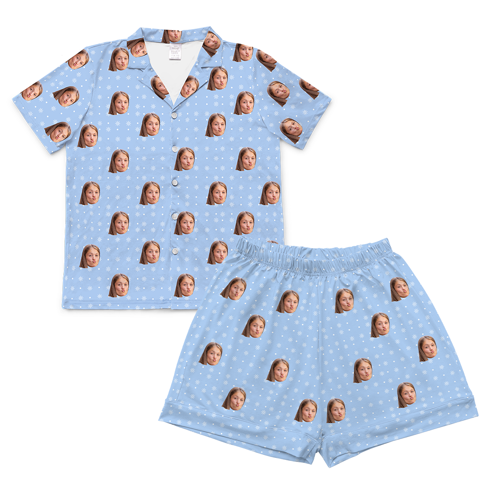 A matching short-sleeved pajama set, consisting of a button-up shirt and shorts. Both pieces feature a light blue background patterned with small white snowflakes. Repeated headshots of a young girl's face, making a playful 'fish face' or blowing a kiss, are superimposed onto the pattern. 
