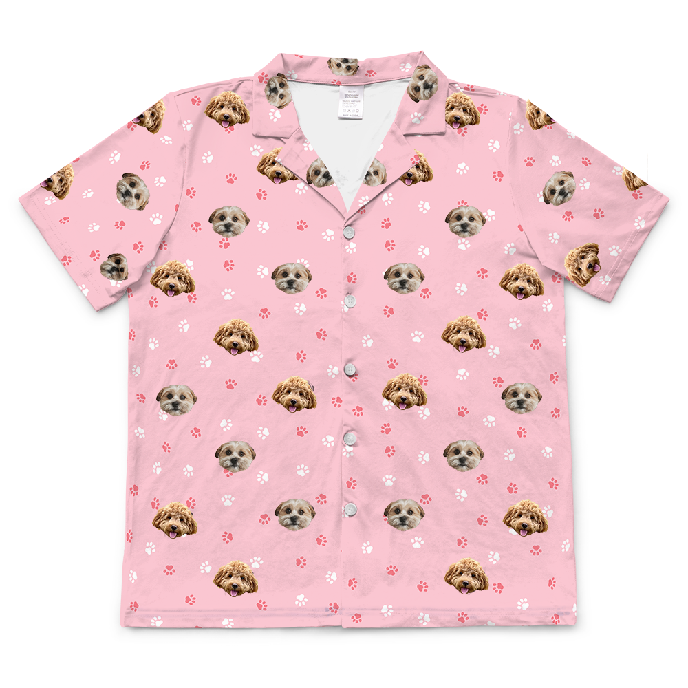 A short-sleeved, button-up pajama top in light pink. The top features a repeating pattern of cartoonish pink and white paw prints, interspersed with realistic photos of two different dogs' heads.