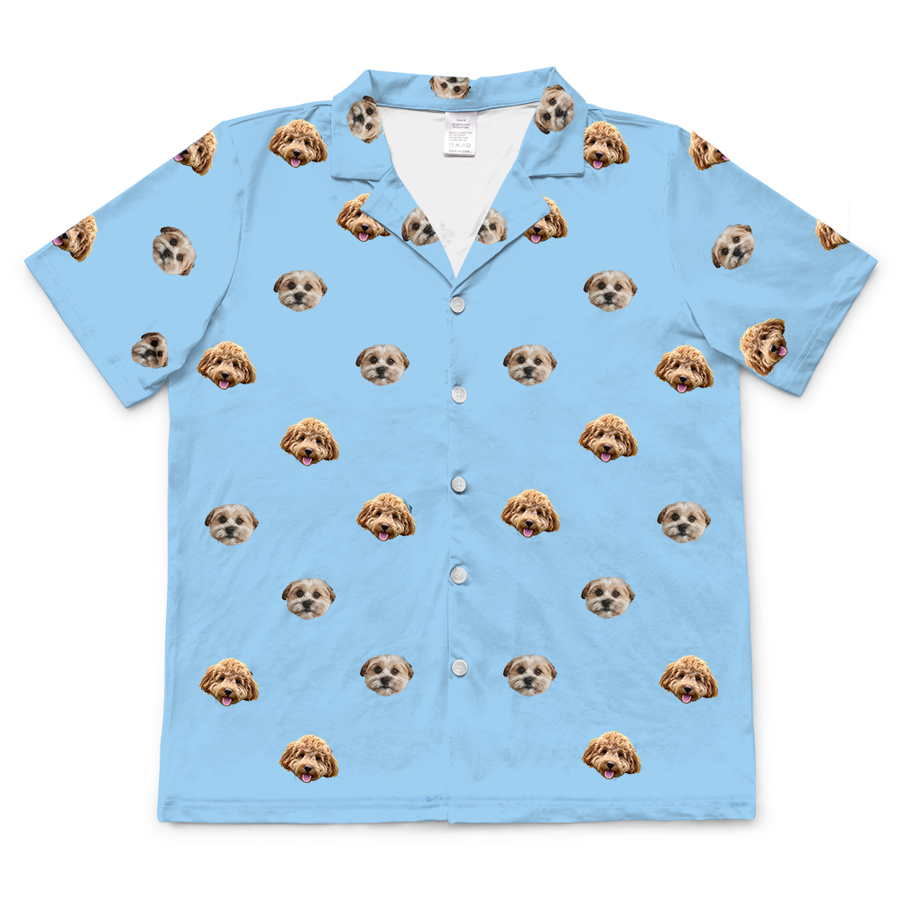 A short-sleeved, button-up pajama top in light sky blue fabric. The top features a repeating pattern of realistic photos of two different dogs' heads.