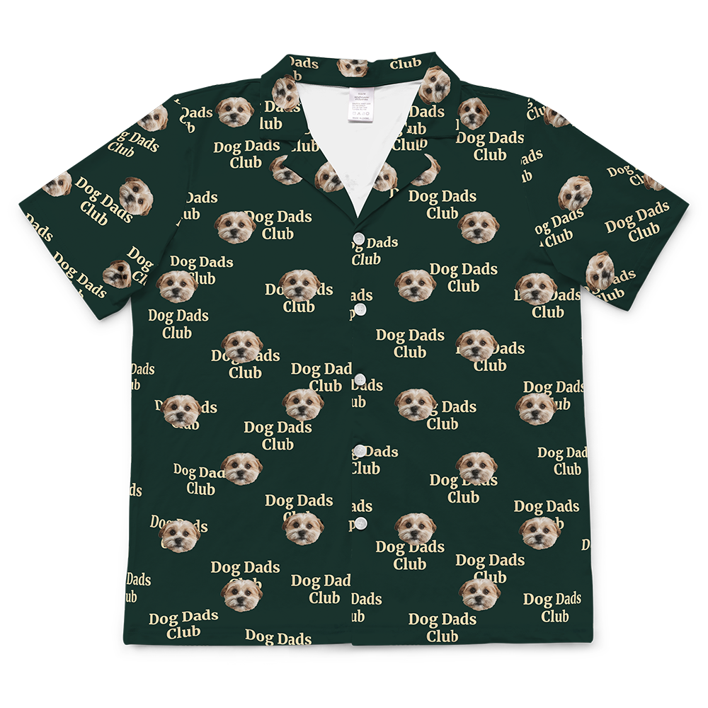 A short-sleeve shirt in a dark green fabric. The shirt is covered in a repeating pattern featuring alternating elements: the phrase "Dog Dads Club" in text, and smiling headshots of a dog.
