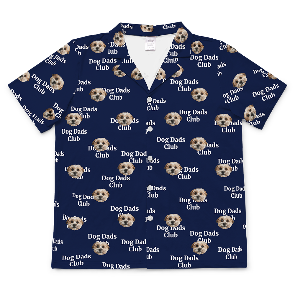 A short-sleeve, button-down pajama top in a dark navy blue fabric. The shirt is covered in a repeating pattern featuring alternating elements: the phrase "Dog Dads Club" in text, and smiling headshots of a dog.