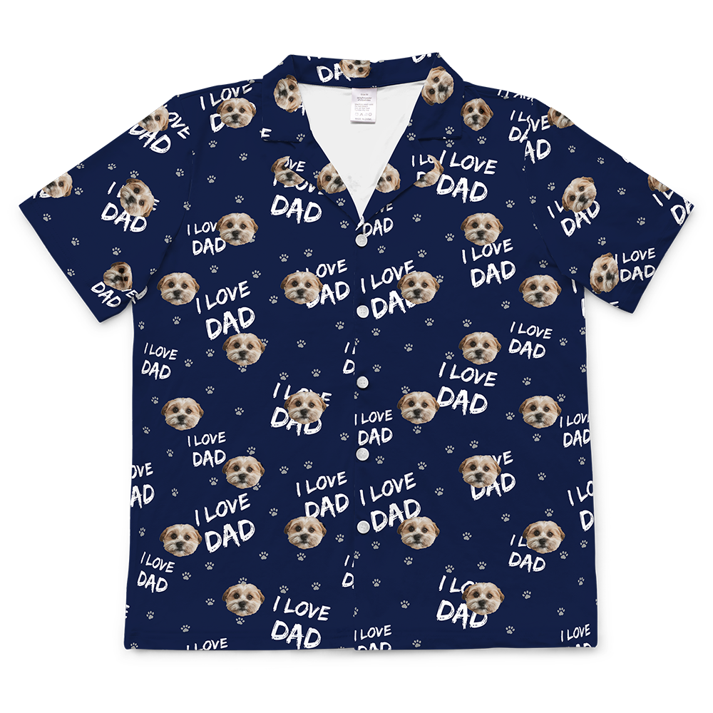 A short-sleeve, button-down pajama top, in a dark navy blue fabric. The shirt is covered in a repeating pattern featuring alternating elements: the phrase "I LOVE DAD" in white text, small  paw print icons, and smiling headshots of a dog.