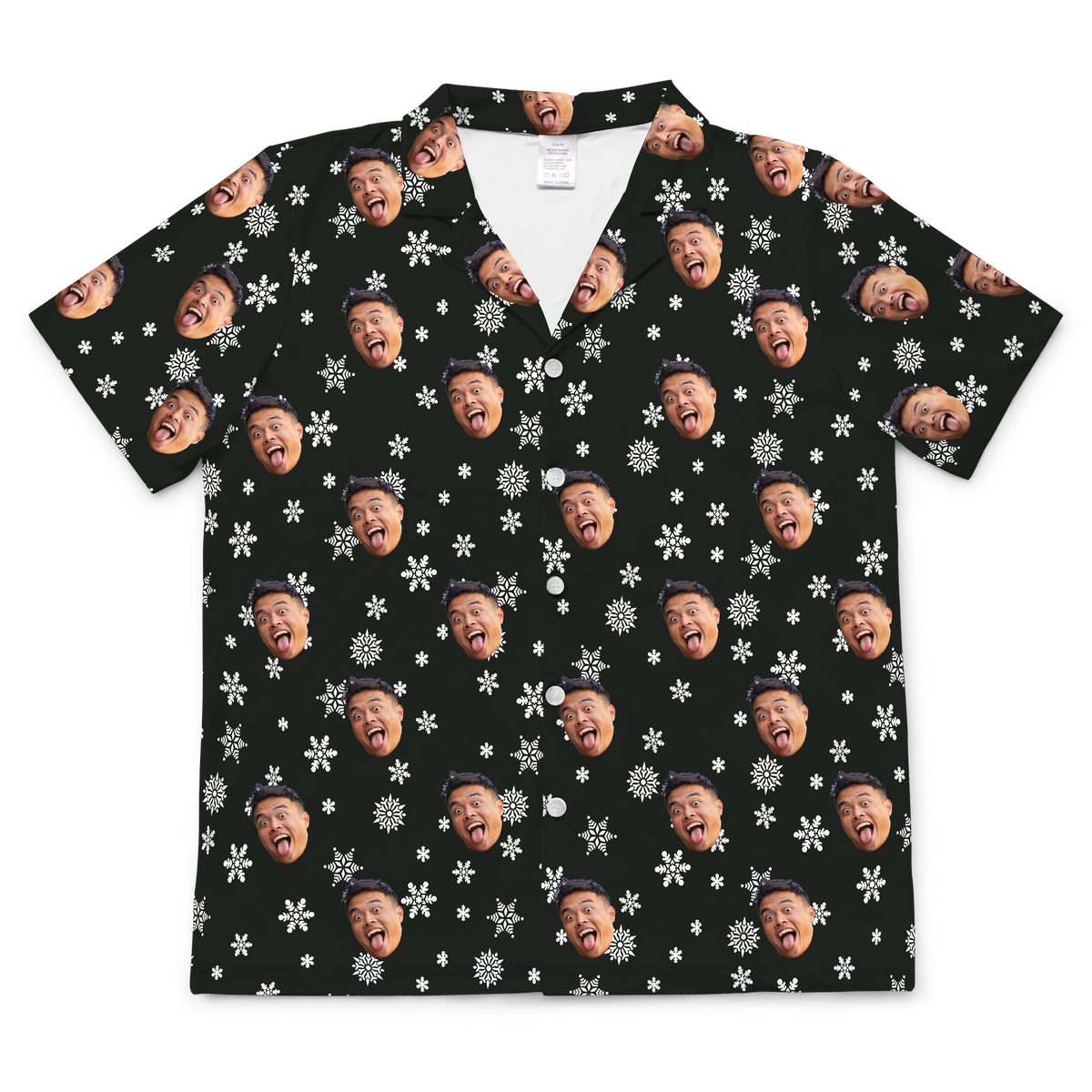 A short-sleeved, button-up pajama or casual shirt featuring a black background patterned with various white snowflakes. Repeated, highly expressive headshots of a person's face, with their mouth wide open and tongue sticking out, are superimposed onto the pattern. 