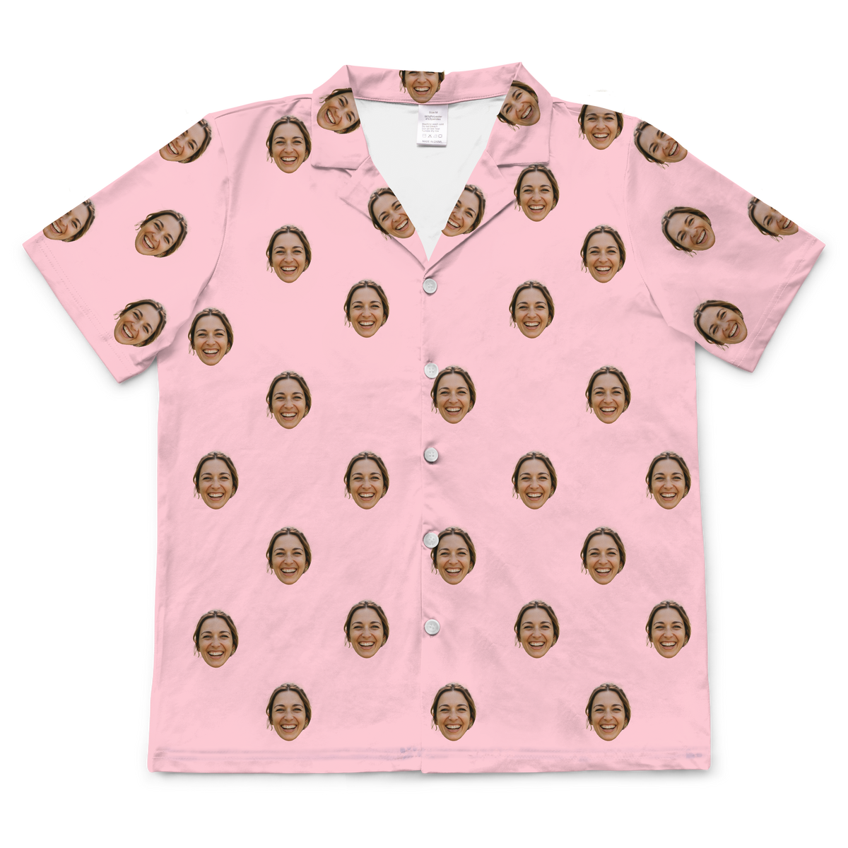A short-sleeved, button-up pajama shirt in a solid light pink or blush-colored fabric. Repeated headshots of a smiling woman's face are evenly spaced and superimposed across the fabric.