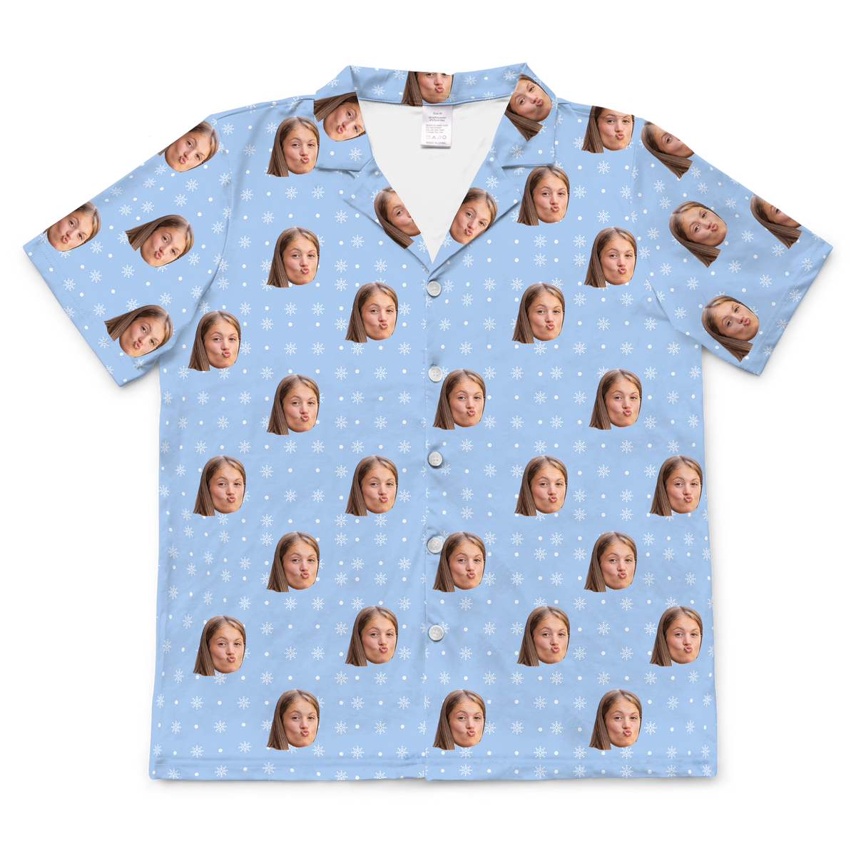 A short-sleeved, button-up pajama or casual shirt featuring a blue background patterned with various white snowflakes. Repeated, highly expressive headshots of a person's face, are superimposed onto the pattern.