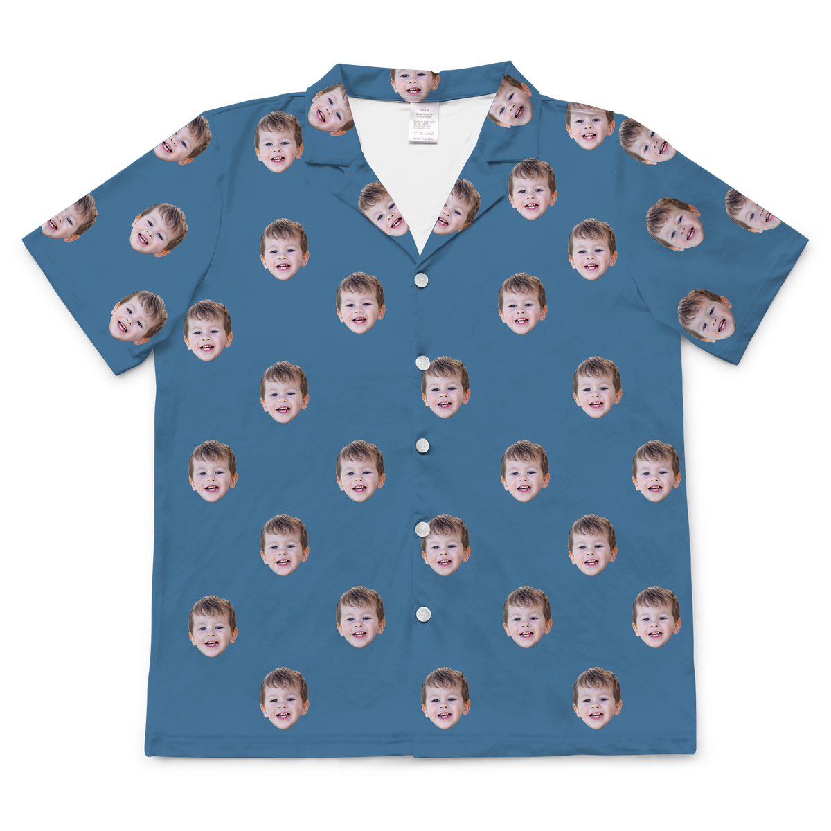 A short-sleeved, button-up pajama or casual shirt in a solid medium blue or denim-colored fabric. Repeated headshots of a young boy's smiling face are evenly spaced and superimposed across the fabric. 