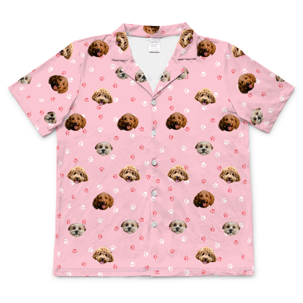 A light pink, short-sleeved, button-down pajama top with a collared neck. It features a repeated pattern of dog faces and paw prints.