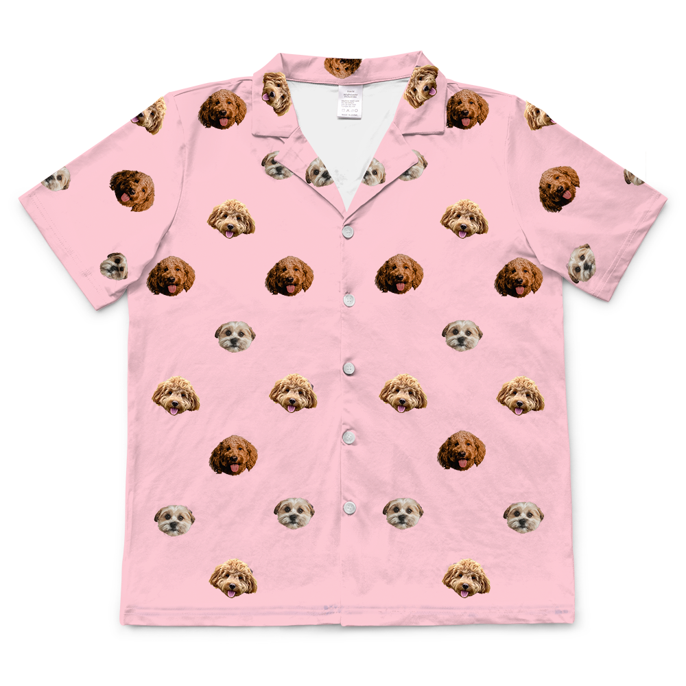 A short-sleeve, button-down pajama top in a light pink color. It features an all-over repeating pattern of the faces of three different small dogs.