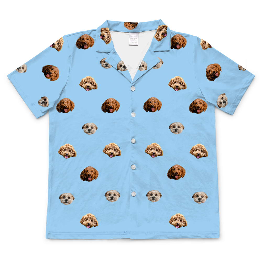 A sky blue, short-sleeved, button-down pajama top with a collared neck. The top features a repeated, scattered pattern of three different dog faces.