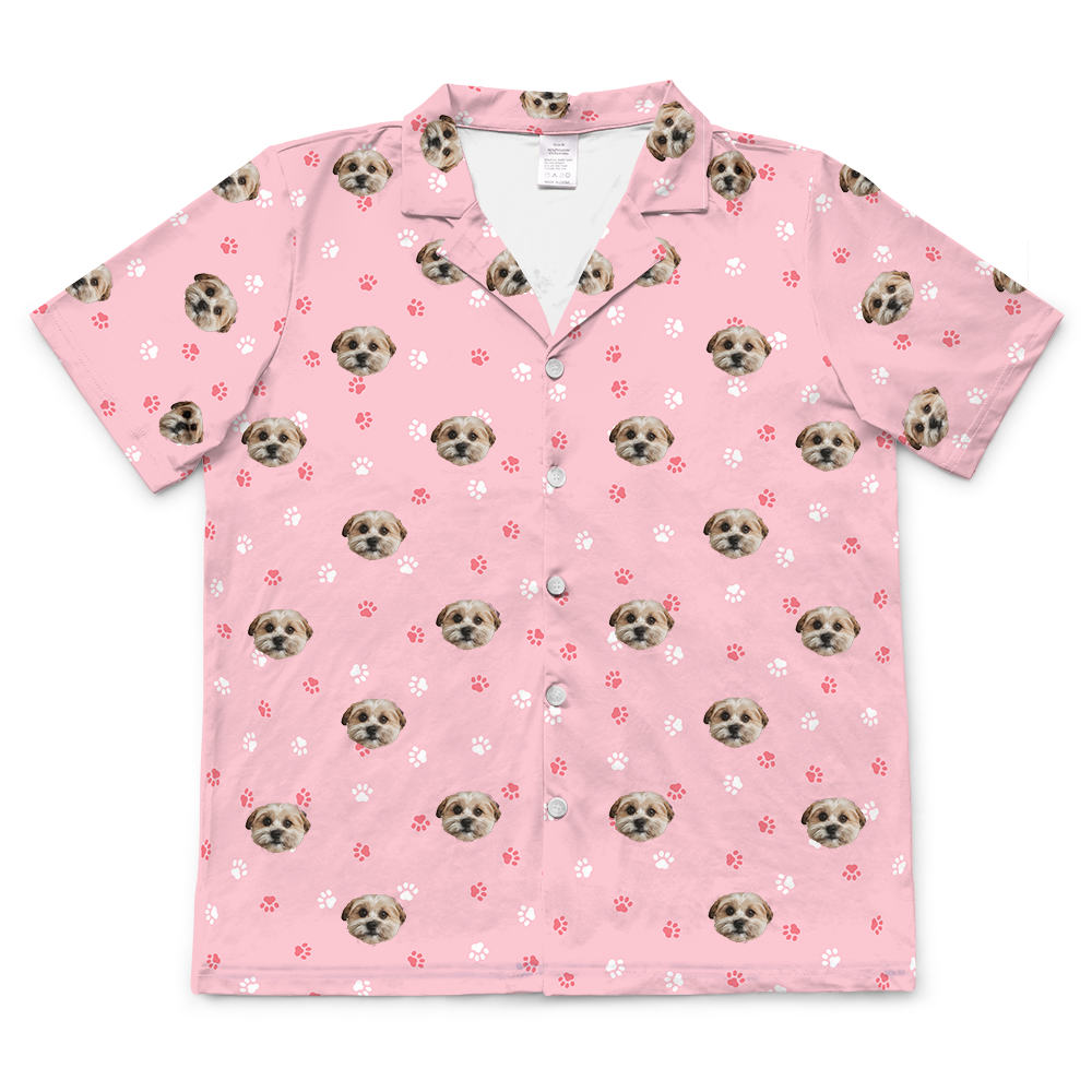 A light pink, short-sleeved, button-down pajama top with a collared neck. The top features a repeated pattern of dog faces.