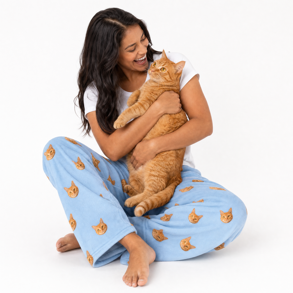 Woman wearing sky blue pajama pants with cat patterns, holding an orange cat on a white background