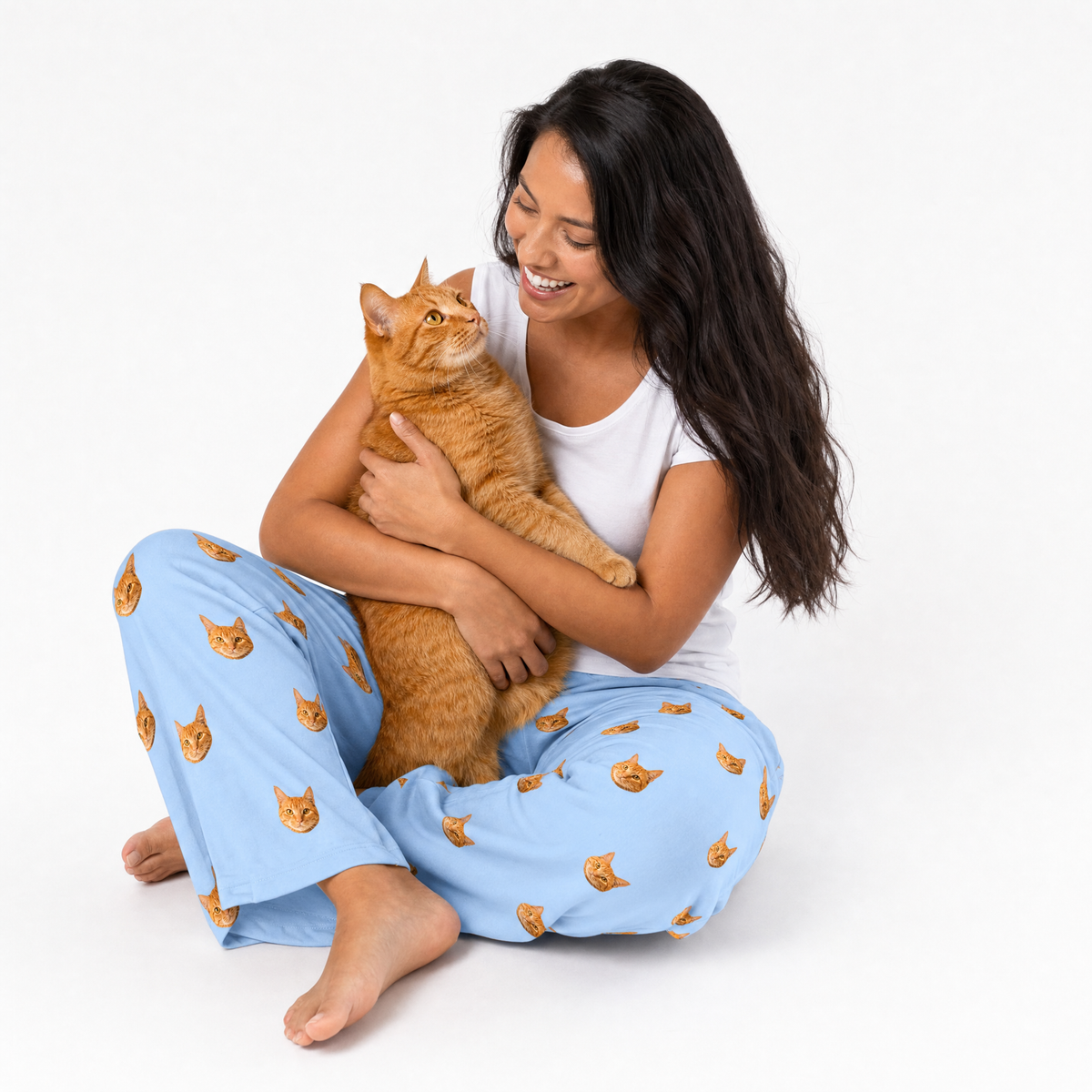 Woman wearing sky blue pajama pants with cat pattern, holding an orange cat on a white background