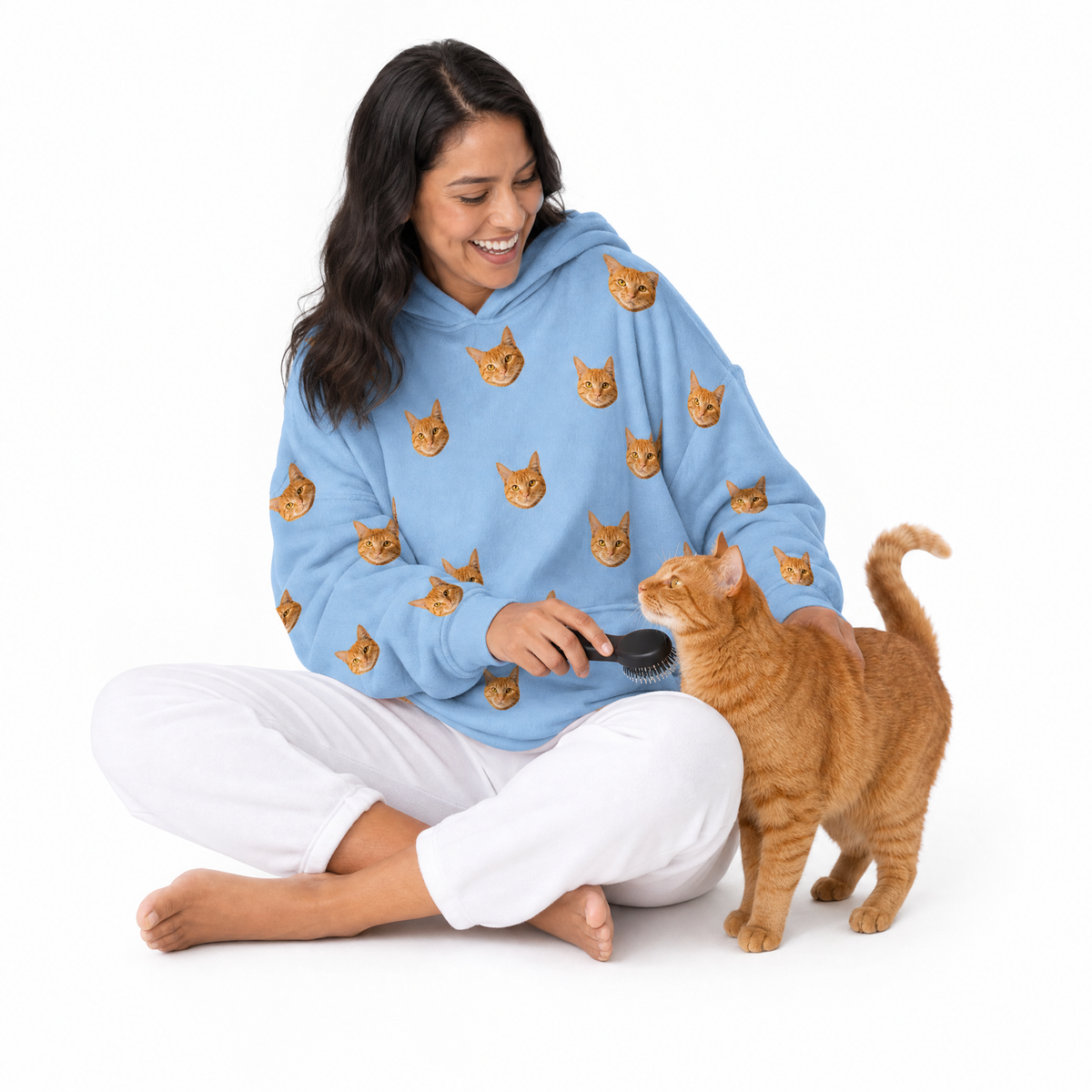 Woman wearing a sky blue blanket hoodie with cat patterns, brushing an orange cat on a white background