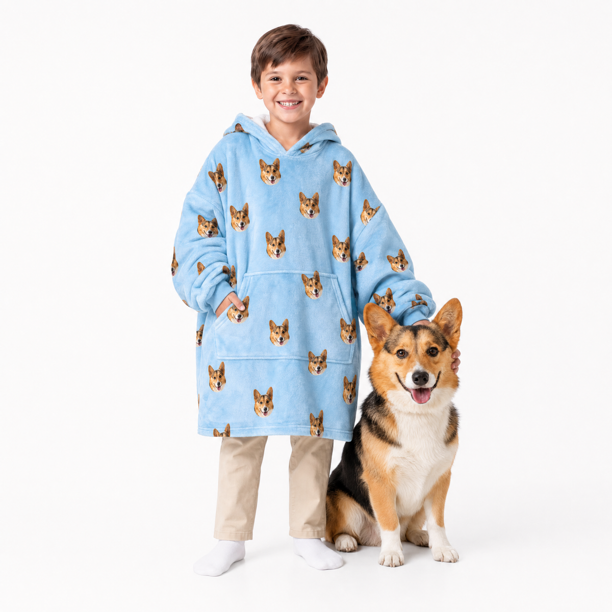 Child wearing a sky blue blanket hoodie with dog pattern standing next to a corgi on a white background