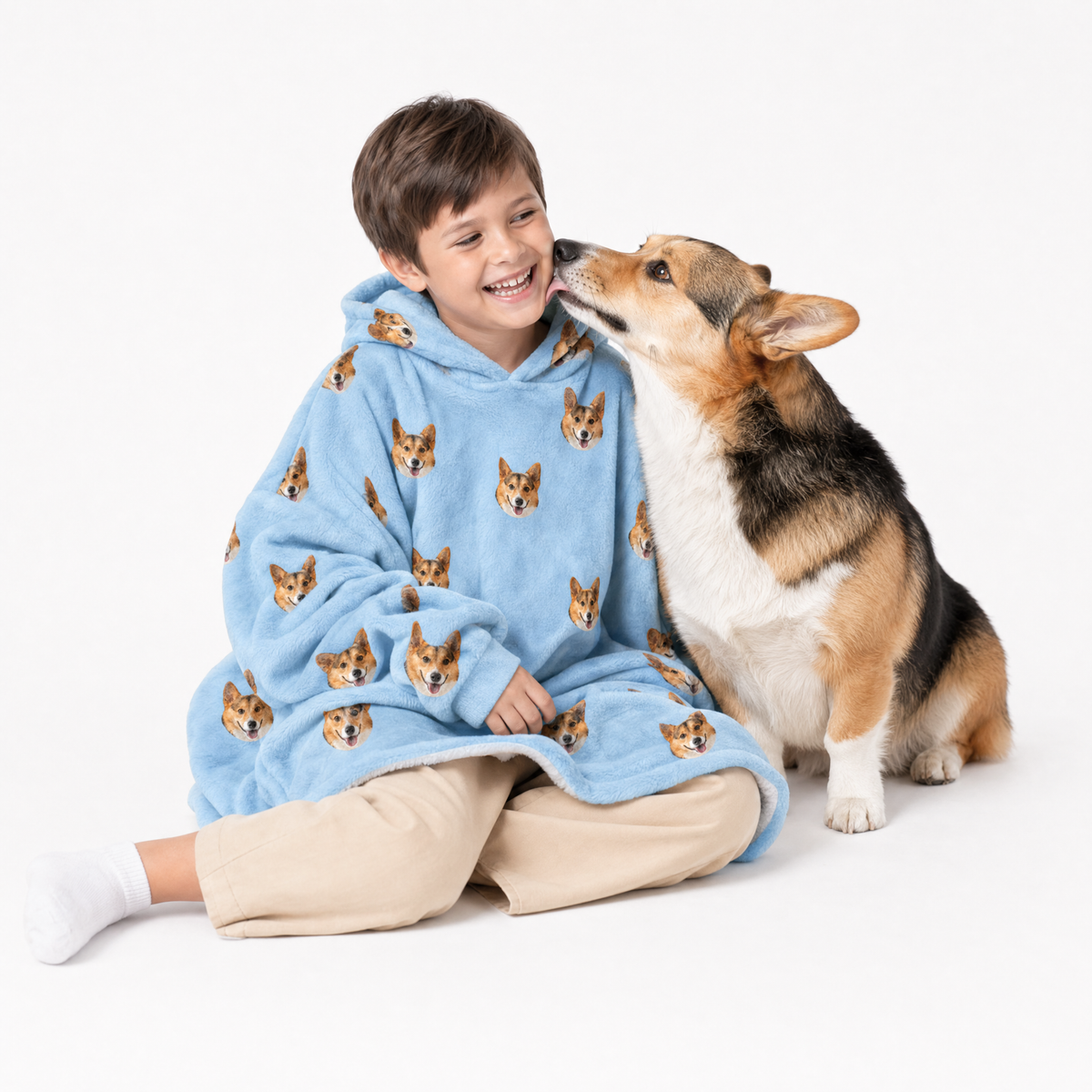 Child wearing a sky blue blanket hoodie with dog patterns, sitting next to a dog on a white background