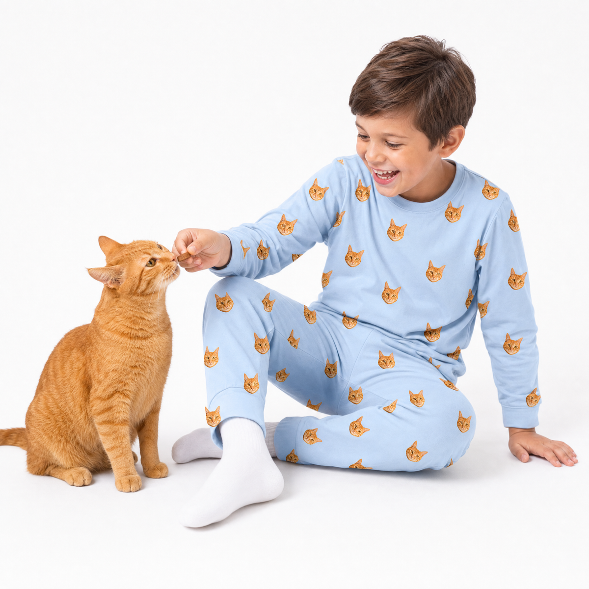 Child in sky blue jogger pajamas with cat pattern feeding a cat on a white background