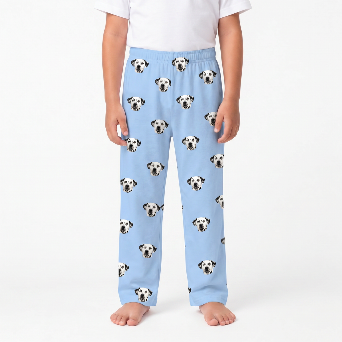 A child wearing sky blue pants with an all-over print of black-and-white dog faces, paired with a plain white top, standing against a light gray studio background.