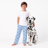 A young boy stands barefoot next to a Dalmatian dog against a plain white studio background. The boy is wearing a white T-shirt and Sky Blue pajama pants covered in a repeating pattern of black-and-white dog faces.
