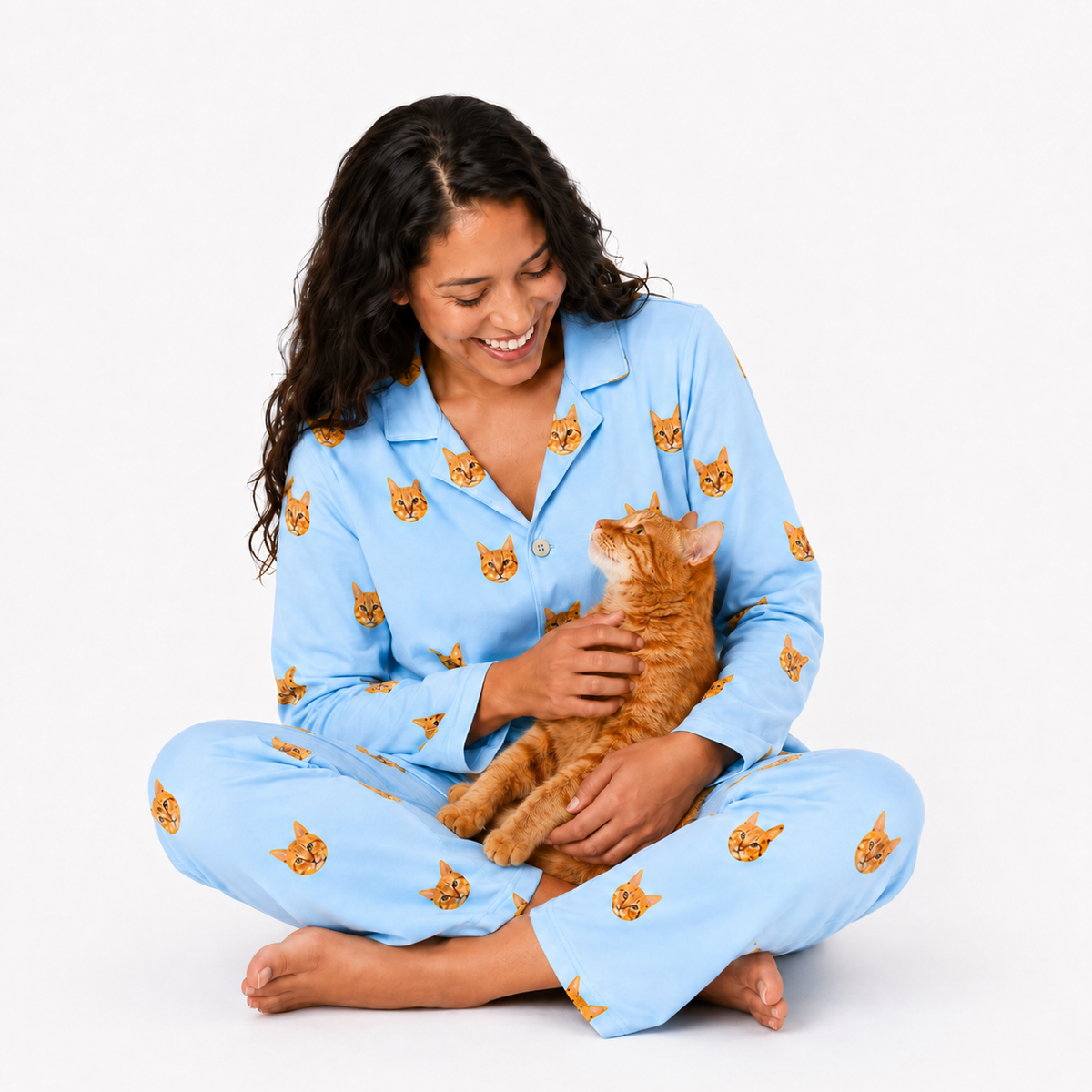 Woman wearing sky blue pajamas with cat patterns, sitting on a white background with a cat.