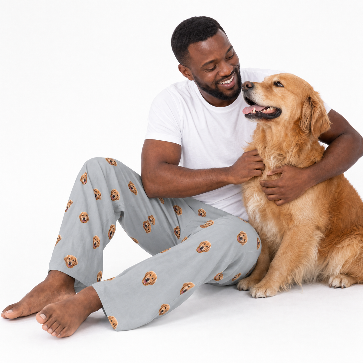 Man wearing pajama pants with dog pattern sitting with a golden retriever on a white background
