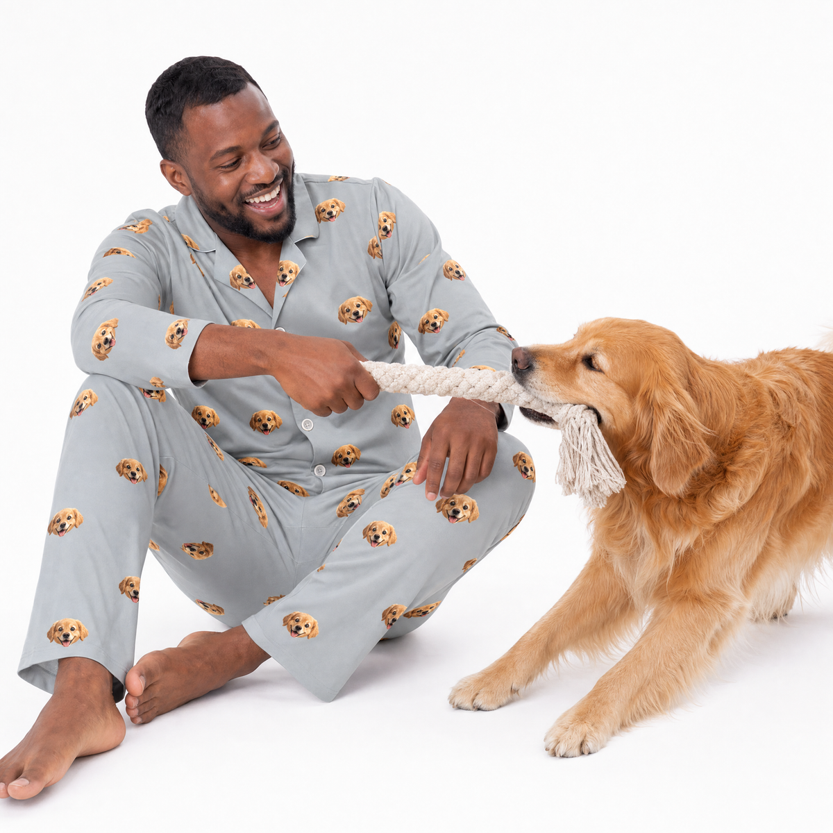 Man wearing a slate gray Golden Retriever print long sleeve pajama set while sitting on the floor and smiling as he plays tug-of-war with a Golden Retriever using a rope toy.