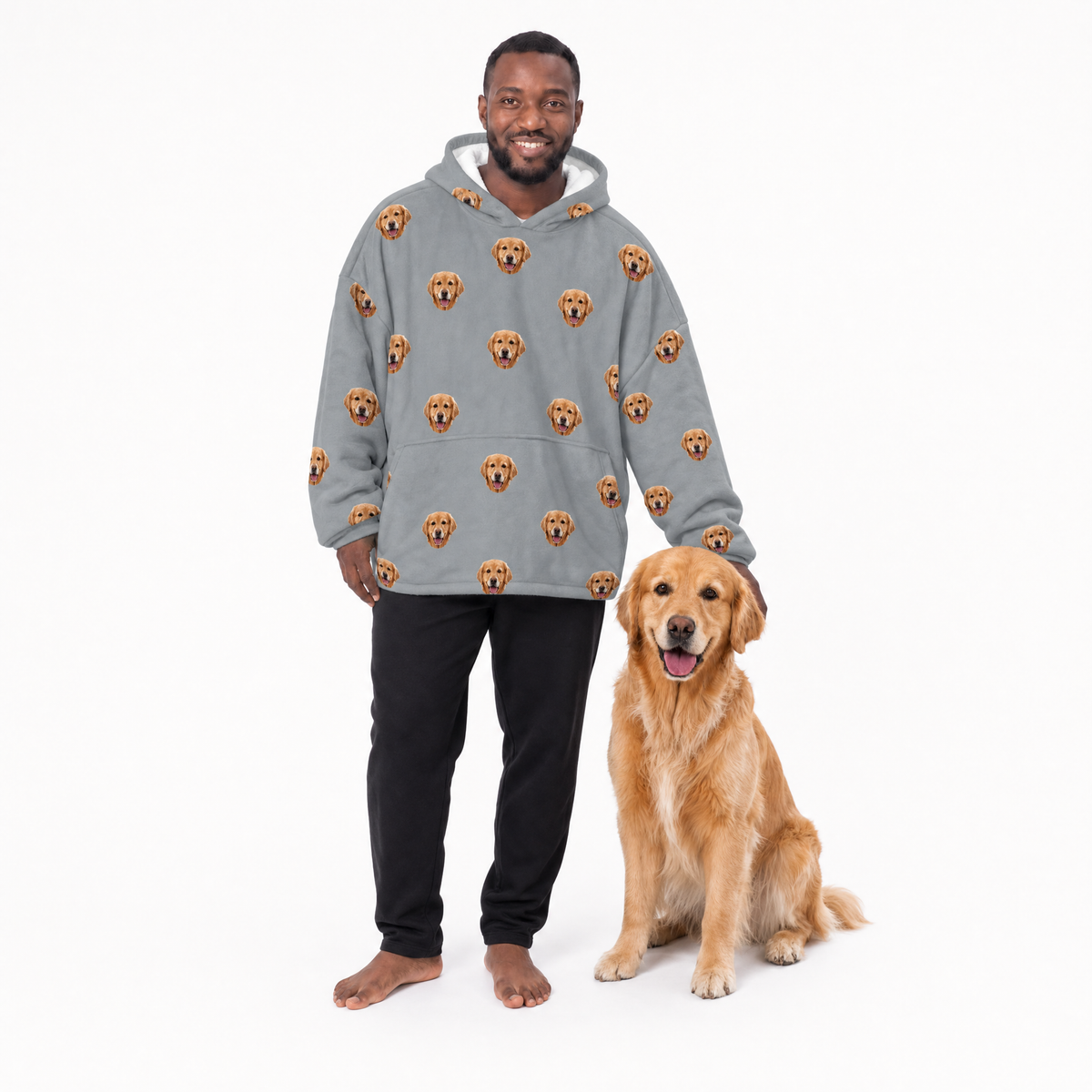 Man wearing a gray blanket hoodie with dog pattern and black pants, sitting next to a golden dog on a white background