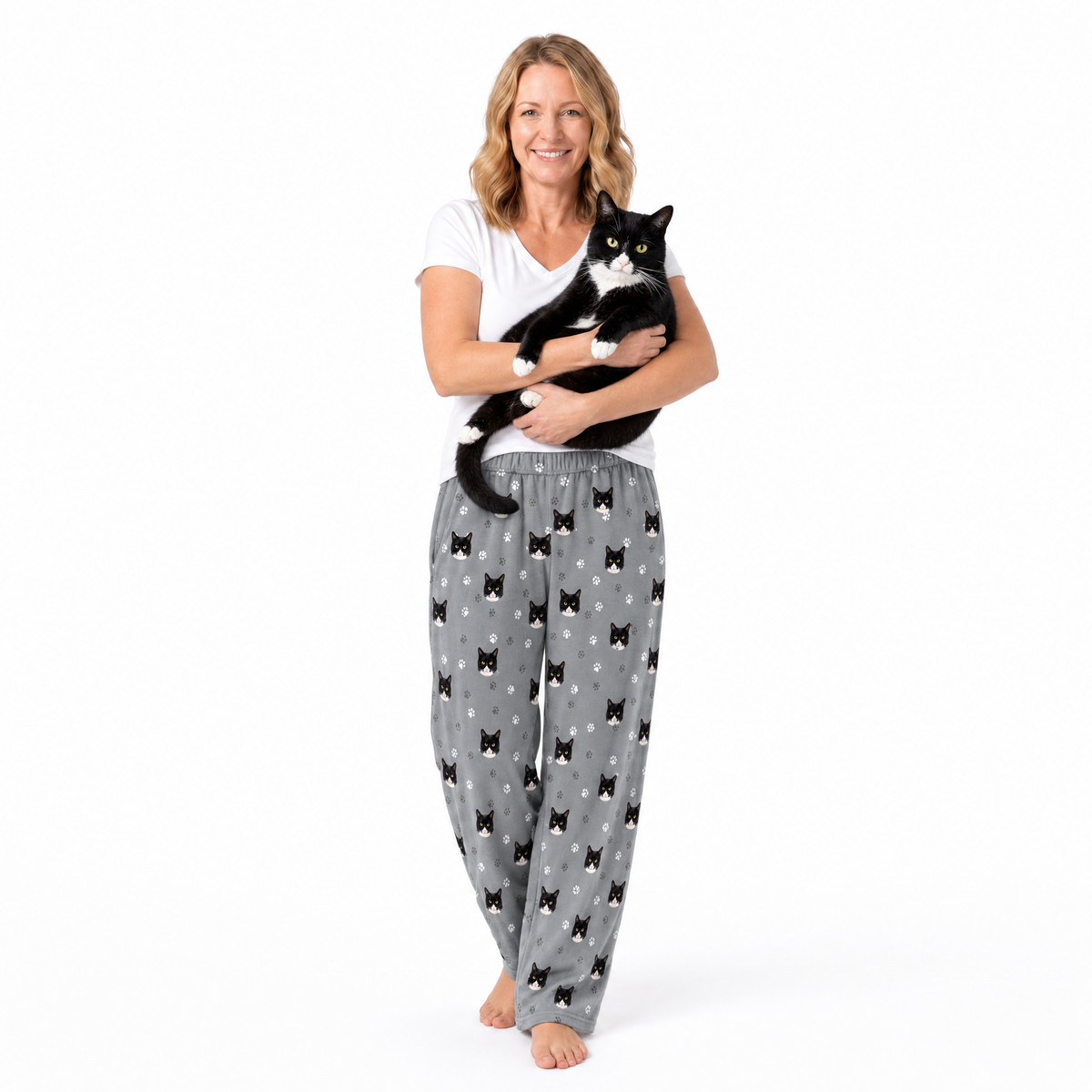Woman wearing gray pajama pants with black cat patterns, holding a black and white cat on a white background