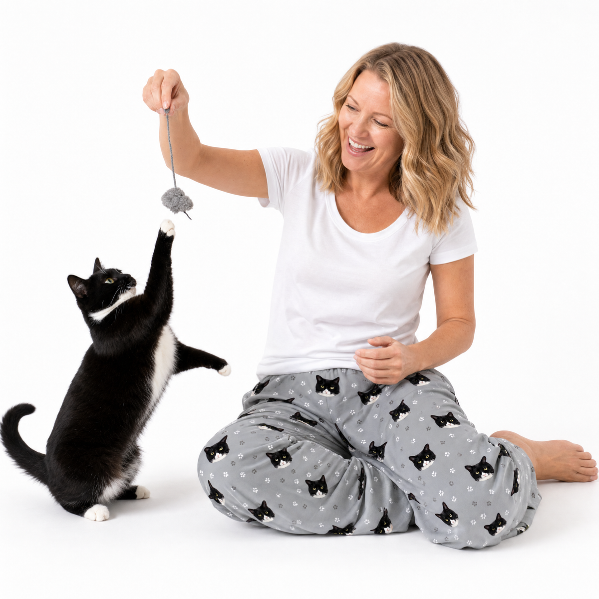 Woman playing with a black and white cat using a toy, both sitting on a white background