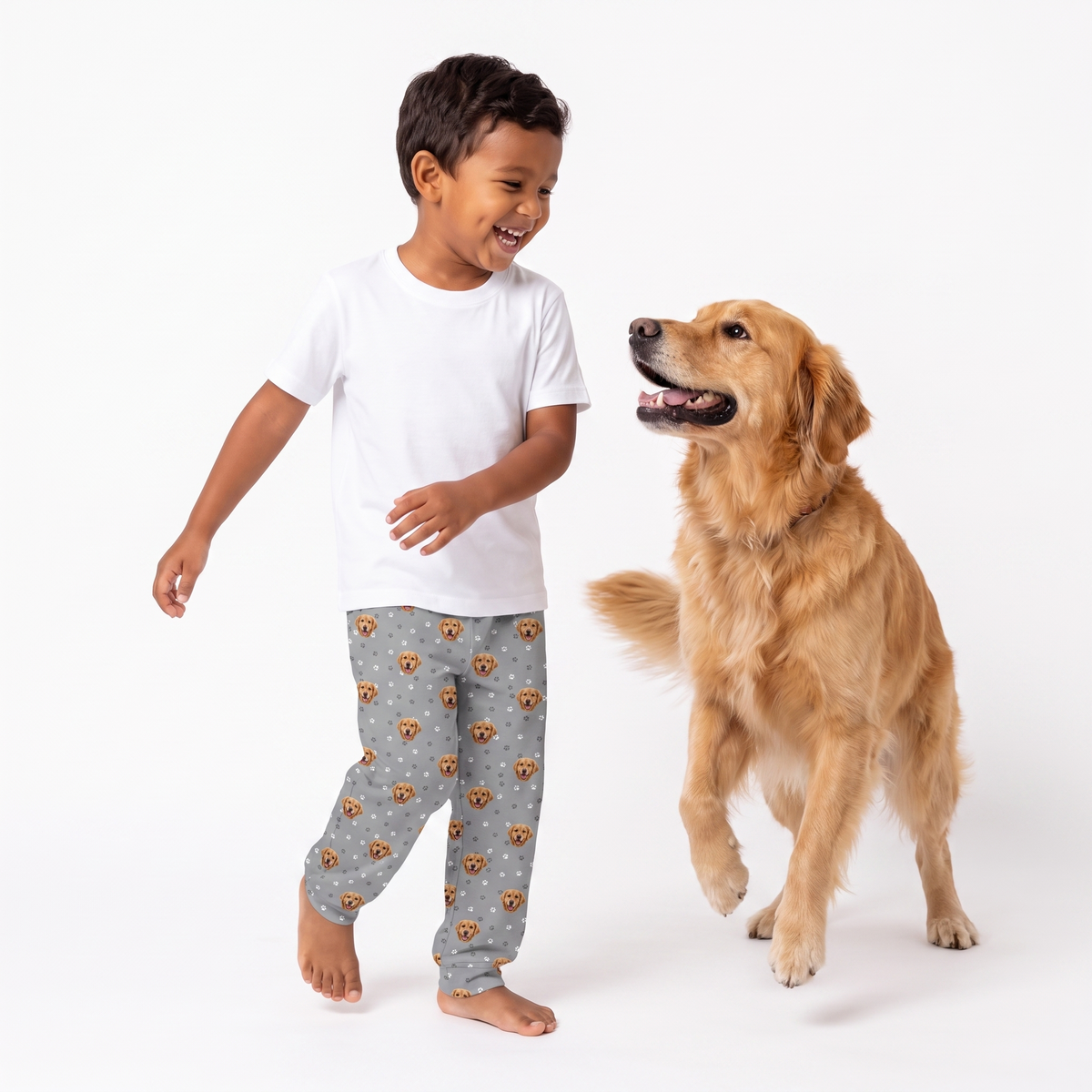 A young boy wearing a white t-shirt and gray patterned pajama pants stands barefoot next to a golden retriever dog on a clean white background.