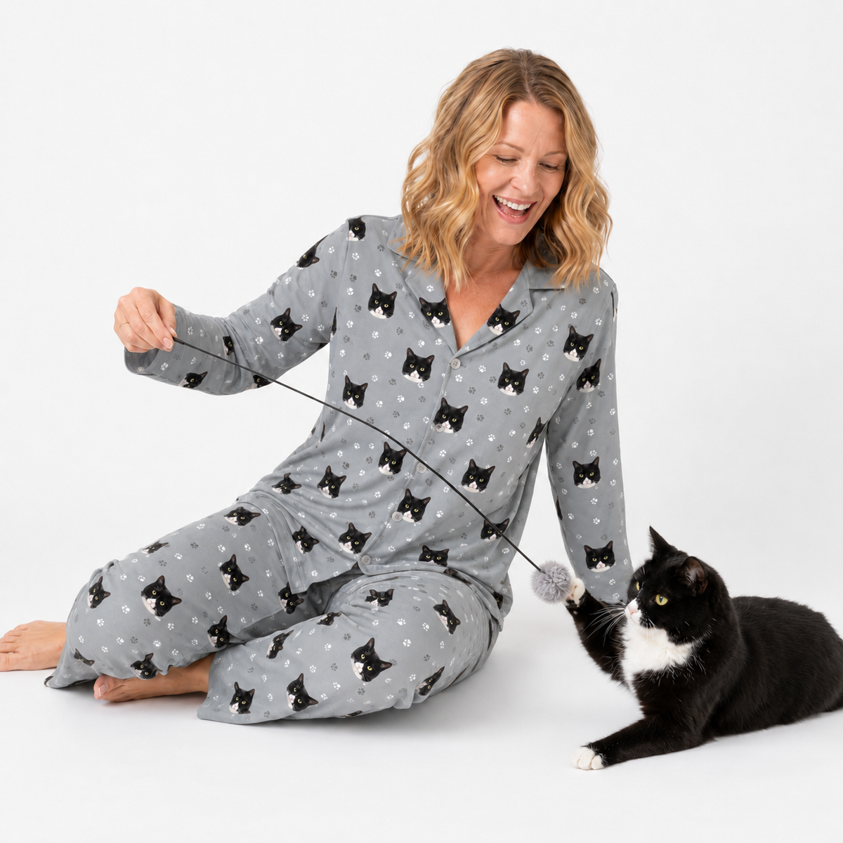 Woman in gray pajamas with black cat pattern sitting next to a black and white cat on a white background