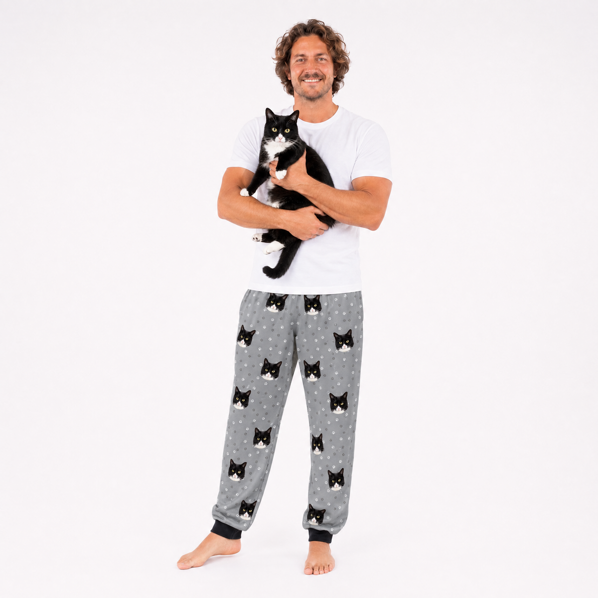 Man wearing a white shirt and gray pants with black cat pattern, holding a black and white cat on a white background.
