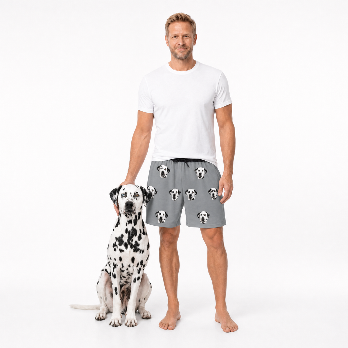 Man wearing a white t-shirt and gray shorts with dog pattern standing next to a Dalmatian dog on a white background