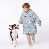 A young boy wearing a light blue hooded blanket jacket with a dog print pattern, walking alongside a black and white dog on a white studio background.