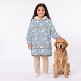 A young girl stands next to a golden retriever dog wearing a light blue hooded sweatshirt covered in a woodland animal print, paired with beige pants and white socks, against a plain white studio background.