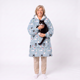 A smiling woman stands against a white studio background wearing a light blue hooded robe with a black-and-white cat print, while holding a black cat. She is also wearing beige pants and white socks.